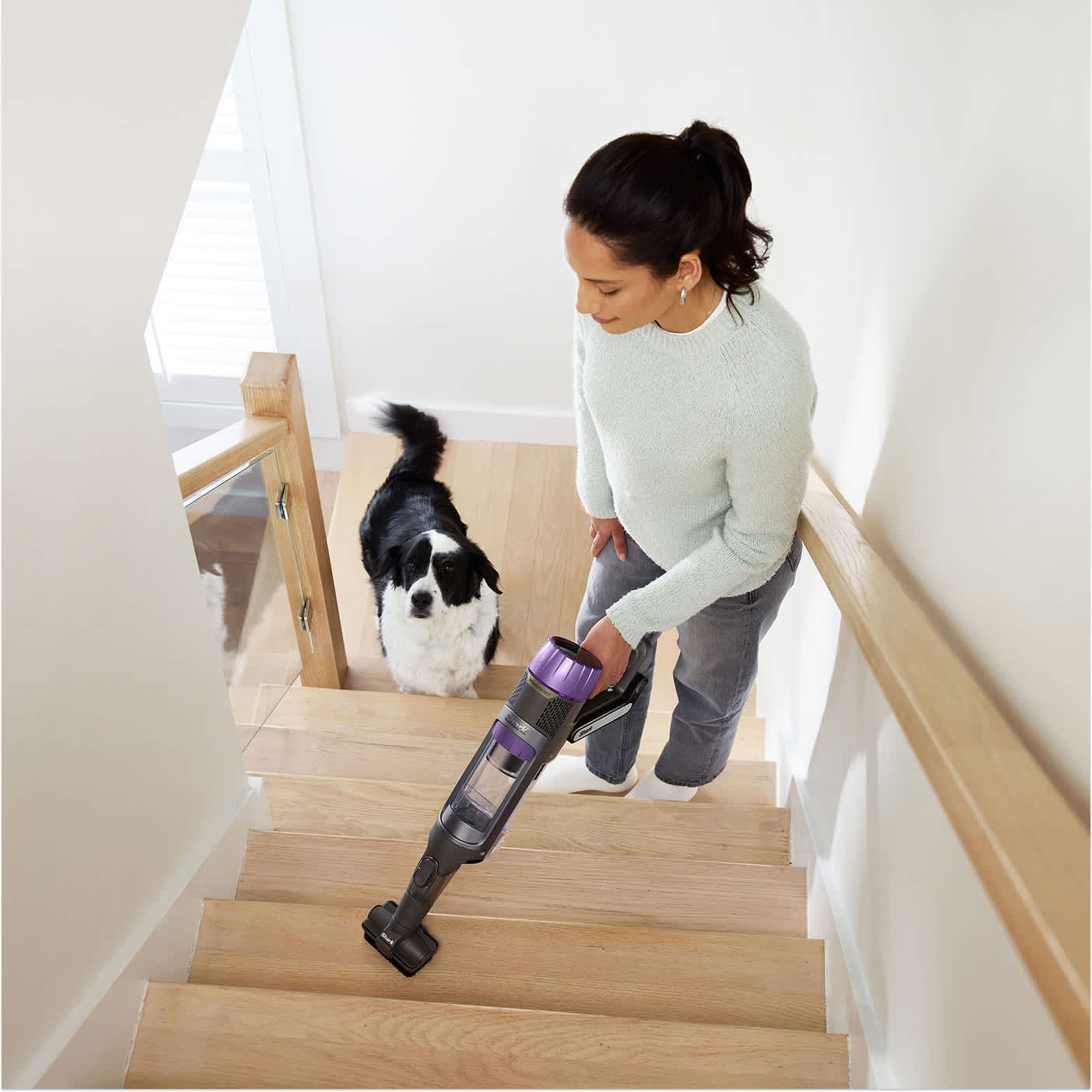 Woman cleaning stairs with the PowerDetect cordless vacuum in handheld mode while a dog stands nearby
